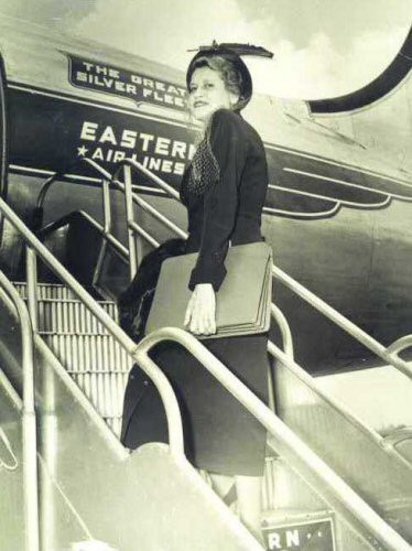 Black and white photo of woman on the stairs of an airplane. She is looking at the camera and holding a case. 