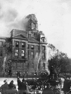 Black and white photograph showing building on fire with people watching in the foreground.