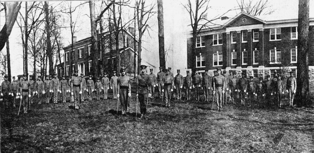 Black and white photograph of a large group of men in military dress standing in front of trees and two brick buildings.