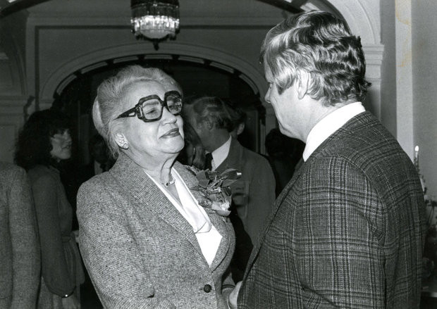 Black and white photo of Mae Street Kidd wearing glasses and talking to a gentleman in plaid suit. 