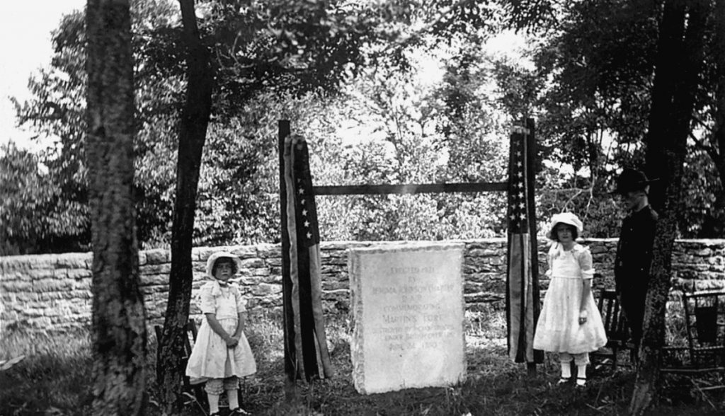 Black and white photograph showing two young girls in white dresses on either side of a stone monument. They are in a wooded area with a stone wall surrounding the stone monument. There is a third individual in the photo who appears to be male and older. 