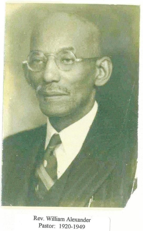 Black and white photograph of older African American man. He is wearing a suit and tie with round rimmed glasses. He is bald with a mustache. 