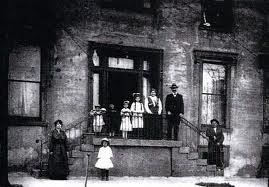 Black and white photo from the 1880s. It shows the entrance to a stone faced building. There are 4 children and 1 adult man and 1 adult woman standing at the stair landing. There is one adult male standing on stairs. There is one child in white standing in front of the landing and 1 adult woman standing to her right. 