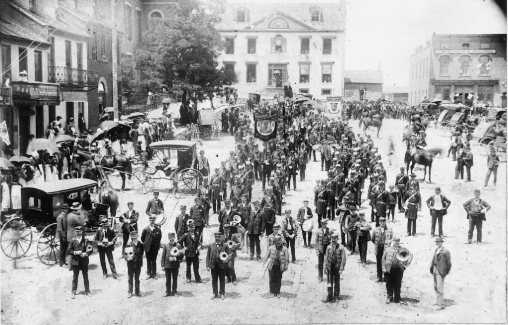 Black and white photograph showing a large group of men standing in lines. Many of the men have musical instruments. There are horses and some cars around the group.